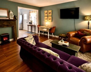 Ein Wohnzimmer mit violettem Sofa, Kamin, Essbereich mit Tisch und Stühlen sowie einem an der Wand befestigten Fernseher.