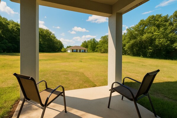 A covered patio with two chairs facing a grassy field and a building in the distance.