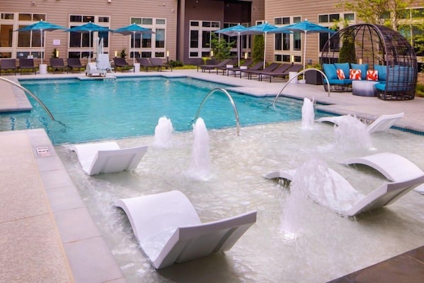 A swimming pool with multiple water jets, lounge chairs, umbrellas, and a seating area with cushions.
