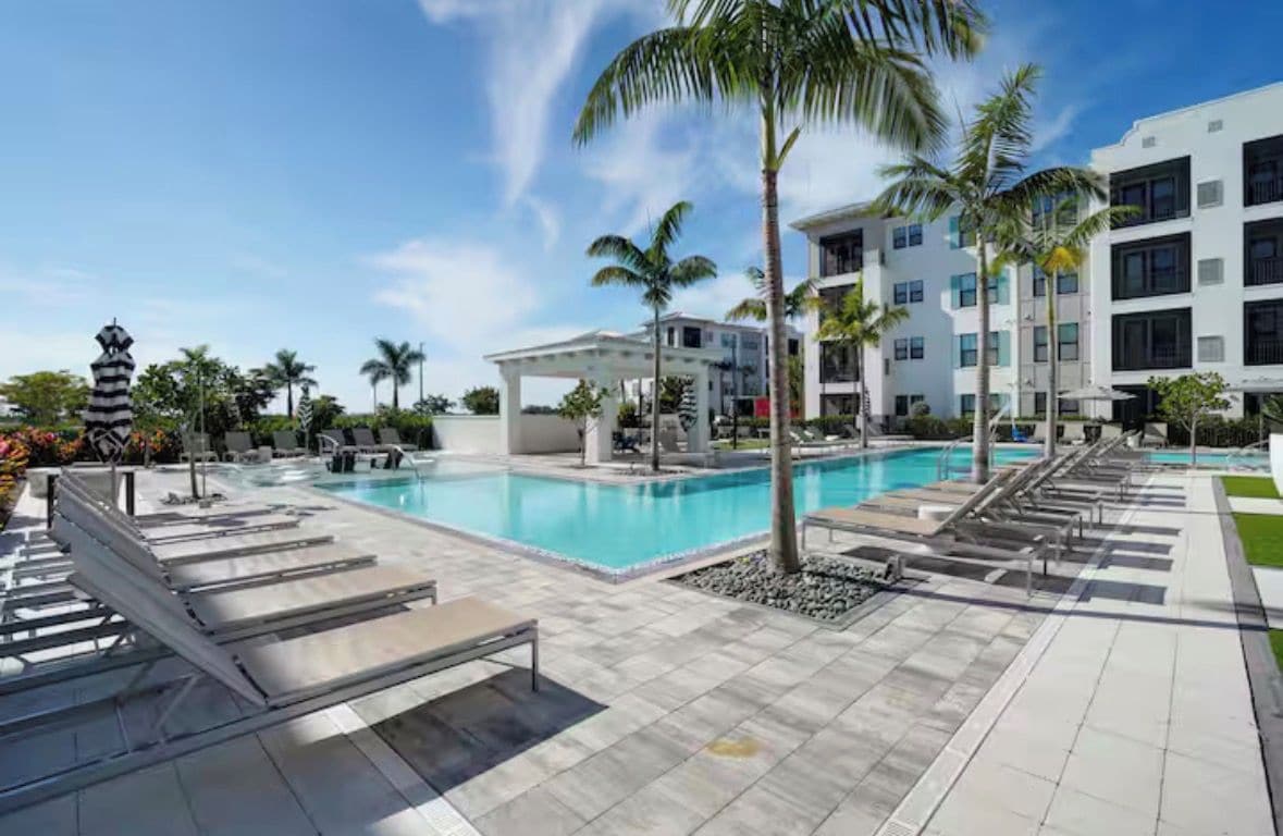 A swimming pool with lounge chairs, palm trees, and a modern building with balconies.
