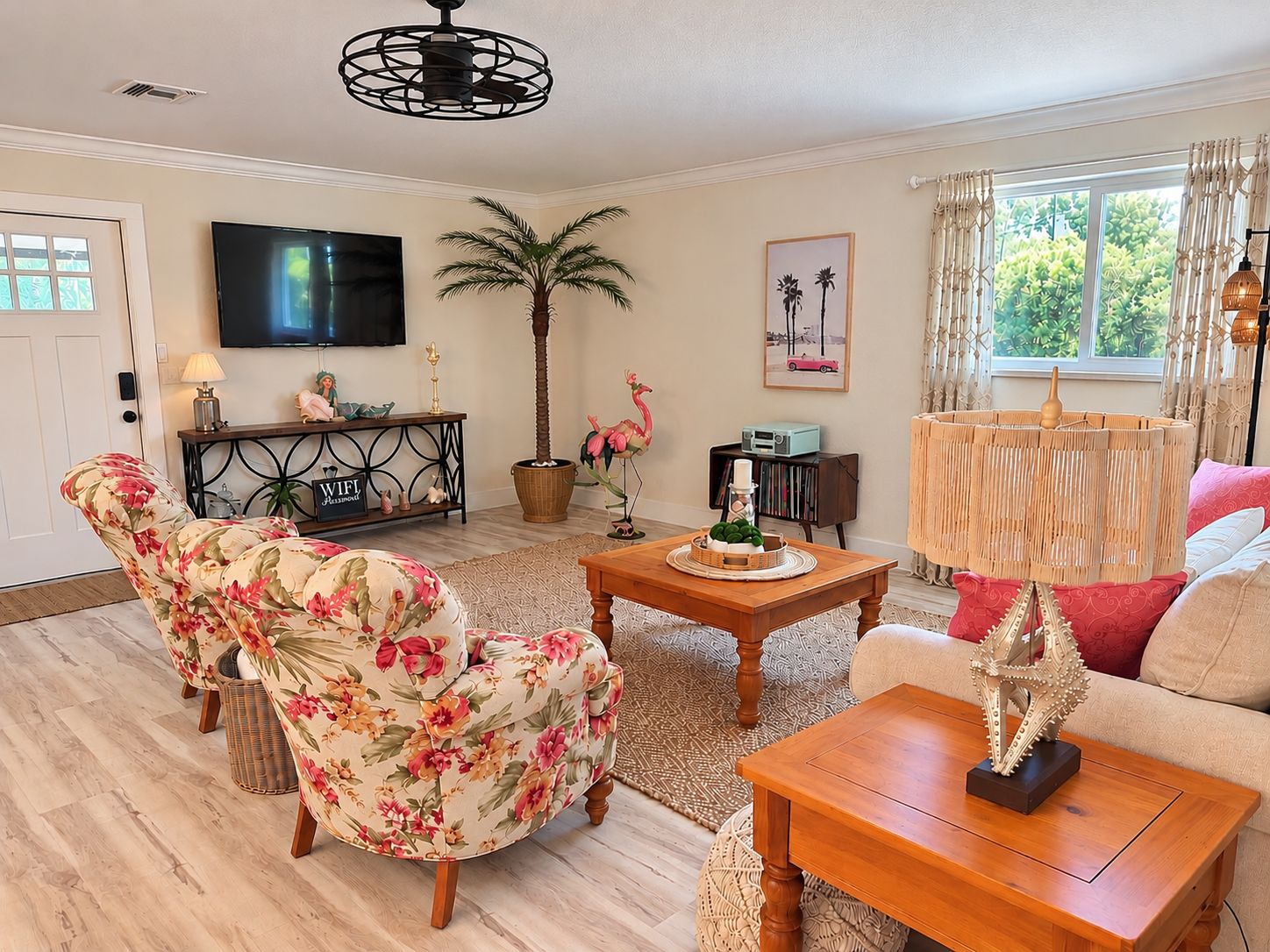 A living room with a flat-screen TV, a wooden coffee table, floral armchairs, a potted palm tree, and a window with curtains.