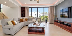 A modern living room with a sectional sofa, a coffee table, a flat-screen TV, and a large window with a view of buildings and greenery. - Villa Almarez | Private Luxury Retreat (Tulum)