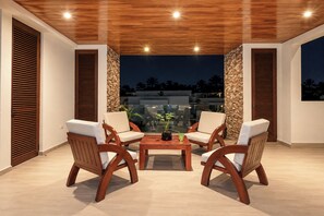 An outdoor patio area with wooden flooring, two wooden chairs with cushions, a small table with a plant, and a view of buildings in the background. - Villa Almarez | Private Luxury Retreat (Tulum)