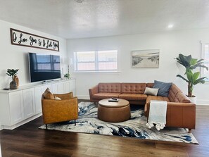 A living room with a sofa, ottoman, and a flat-screen TV. - Furnished 5BR/3BA Home in Bayshore Beautiful — 30-Day Minimum (Tampa)