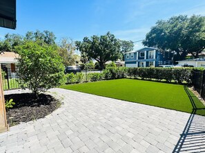 A paved driveway leading to a well-maintained lawn with a large tree and a blue building in the background. - Furnished 5BR/3BA Home in Bayshore Beautiful — 30-Day Minimum (Tampa)