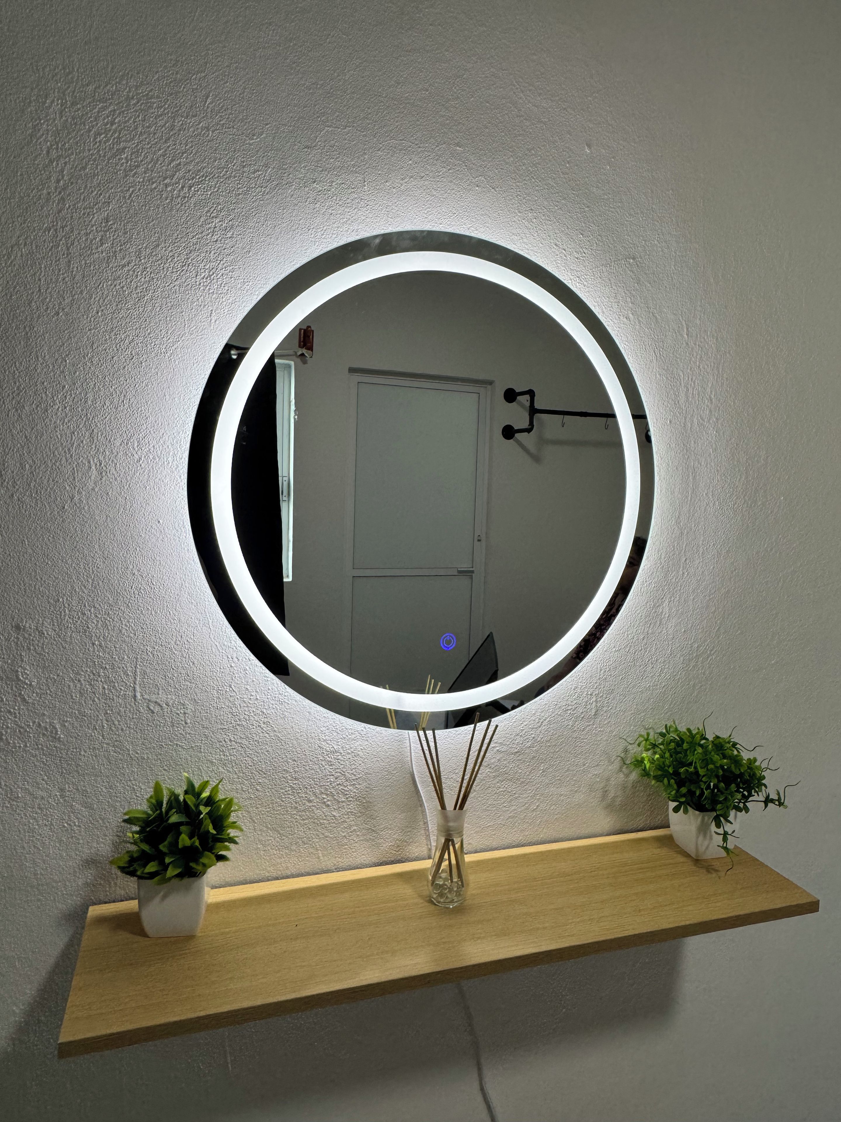 A round mirror with a light halo effect, mounted on a textured wall above a wooden shelf with two potted plants.