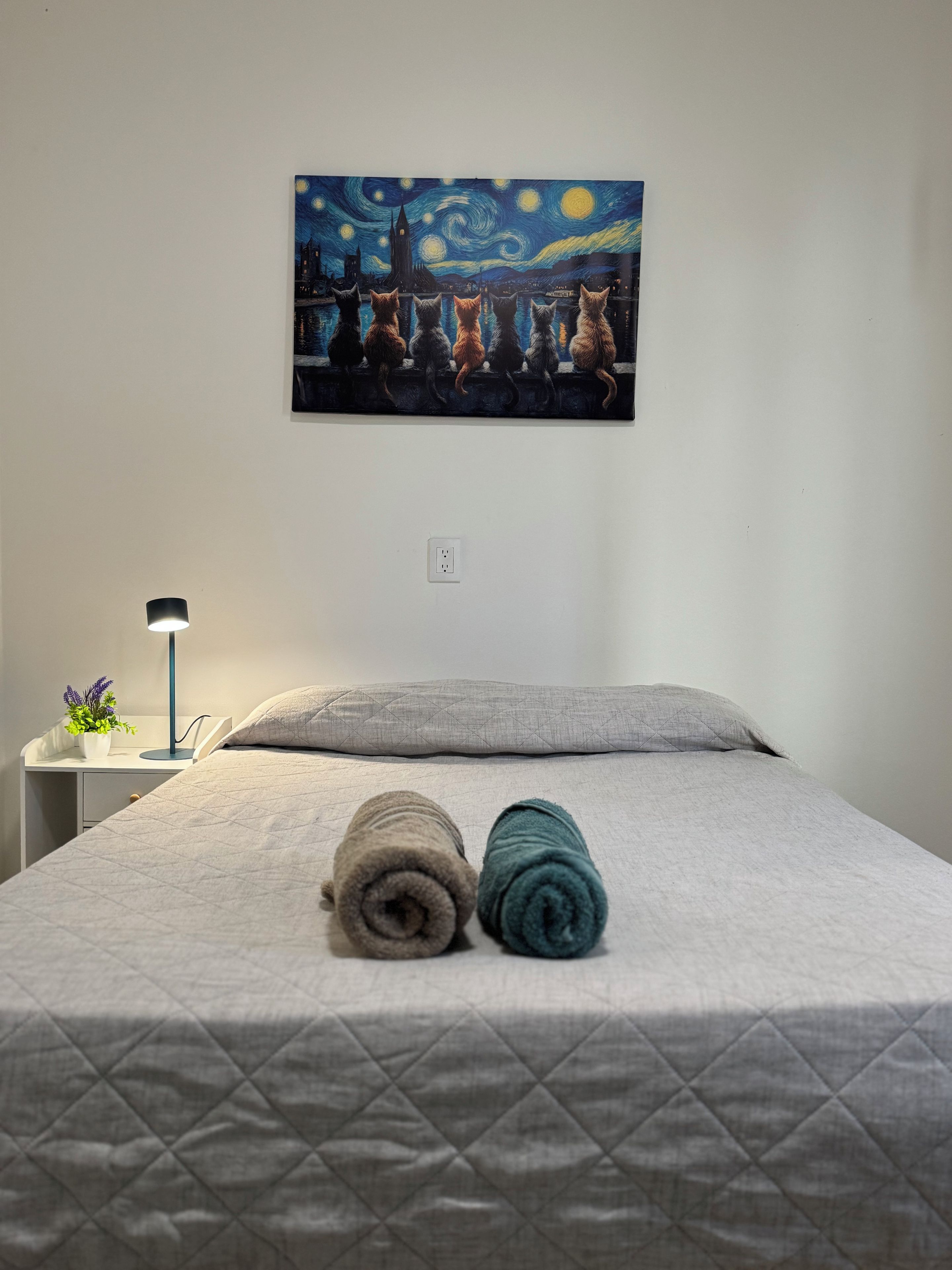 Una habitación de hotel con una cama, un cuadro de una escena nocturna, una mesita de noche con una lámpara y un enchufe en la pared.