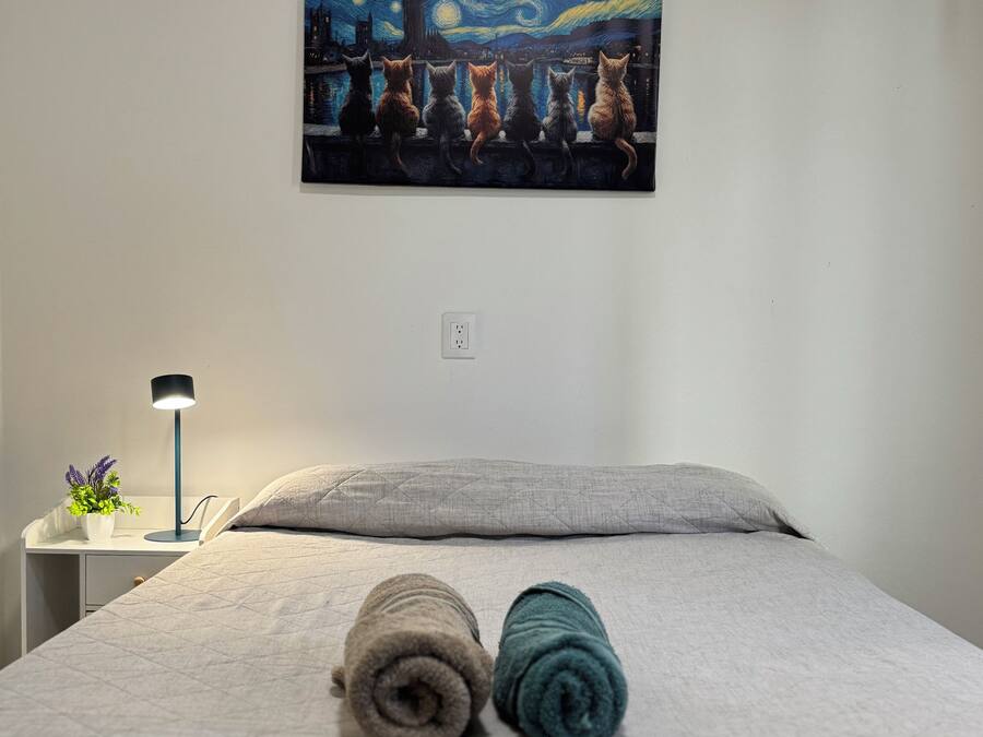 Una habitación de hotel con una cama, un cuadro de una escena nocturna, una mesita de noche con una lámpara y un enchufe en la pared.