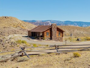 A rustic log cabin with a stone chimney, surrounded by a dry, hilly landscape. - The Lodge at Wapiti Valley Ranch- ideally located between Cody and Yellowstone (Cody)