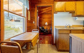 Interior de una cabaña de madera con una mesa de comedor, sillas, una cocina con lavamanos y vistas a través de una ventana.