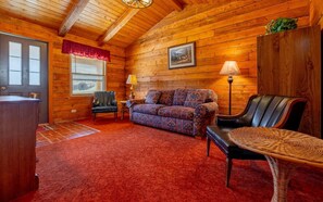 A log cabin interior with a red carpet, a patterned sofa, a leather chair, a wooden cabinet, and a framed picture on the wall. - The Cabin at Wapiti Valley Ranch- ideally located between Cody and Yellowstone (Cody)