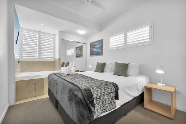 A bedroom with a large bed, a bedside table with a lamp, a built-in bathtub, and a window with blinds. - Studio Apartment - Poolside Tower - No Balcony (Kings Beach)