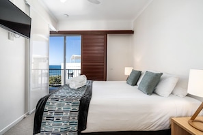 A hotel room with a bed, a bedside table, a TV, and a view of the sea through a sliding door. - 2 Bedroom Rooftop Poolside (Kings Beach)