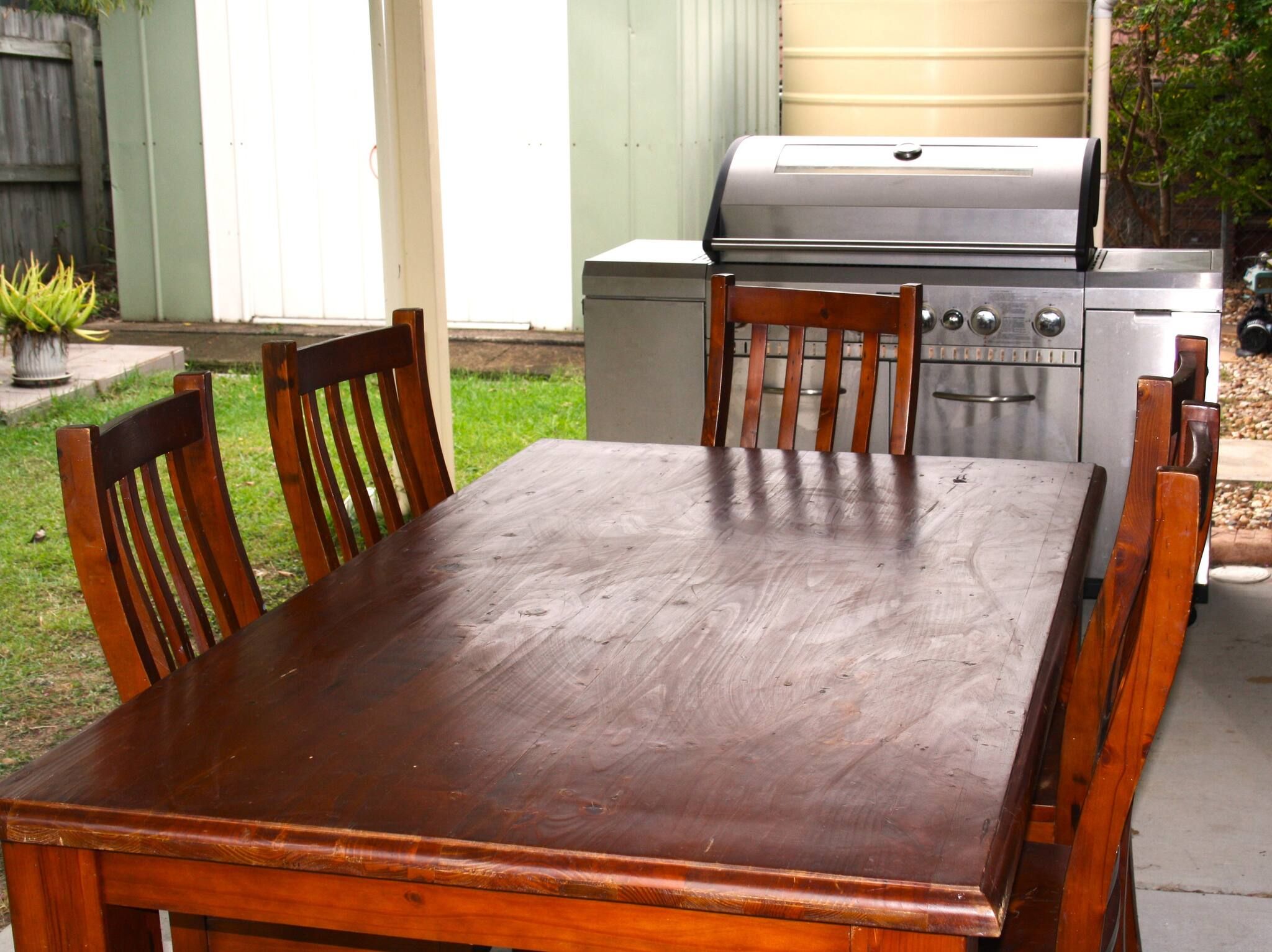 A wooden dining table with chairs, a barbecue grill, and a water tank in the background.
