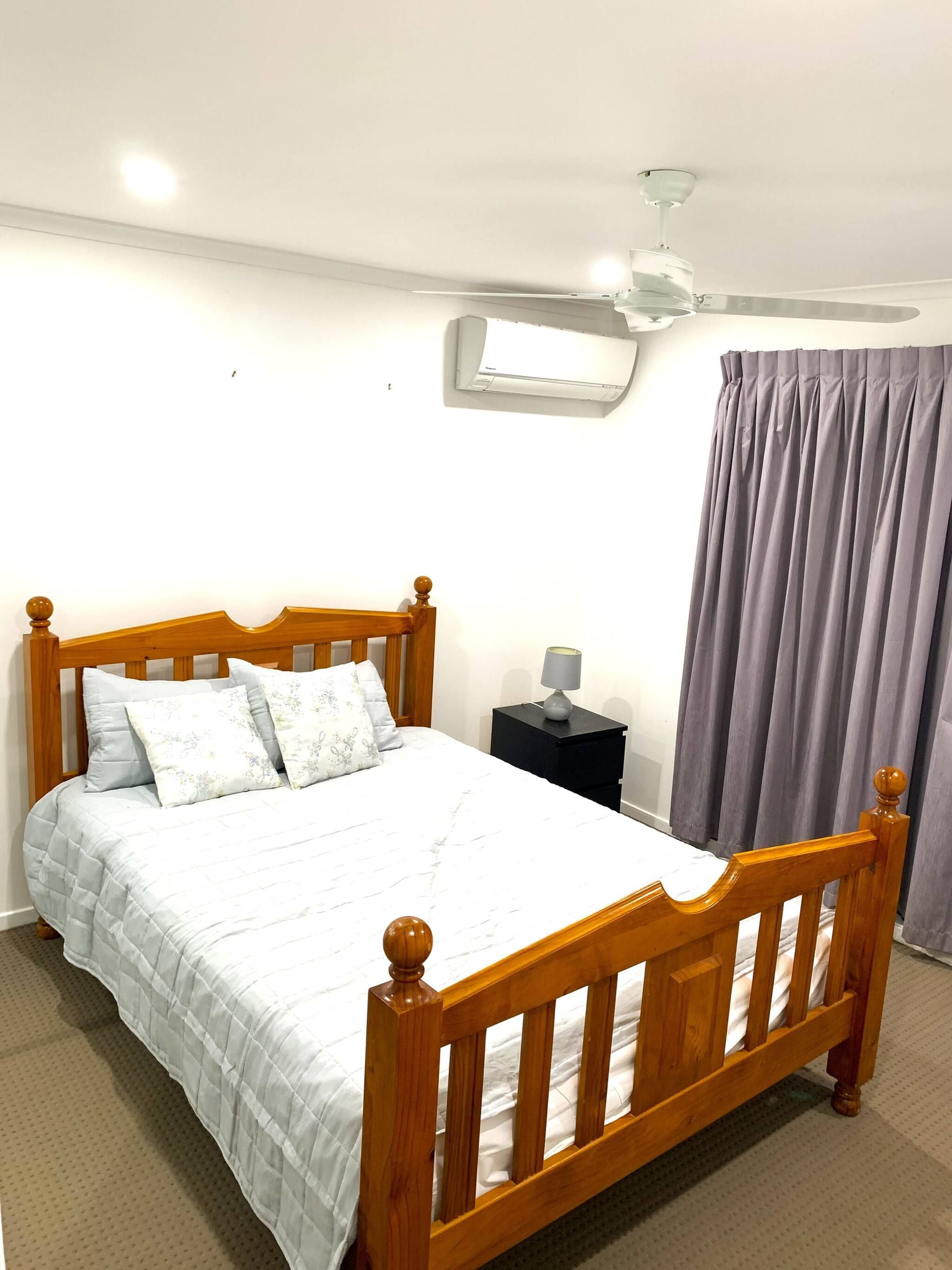 A bedroom with a wooden bed, a nightstand with a lamp, a ceiling fan, and a window with curtains.