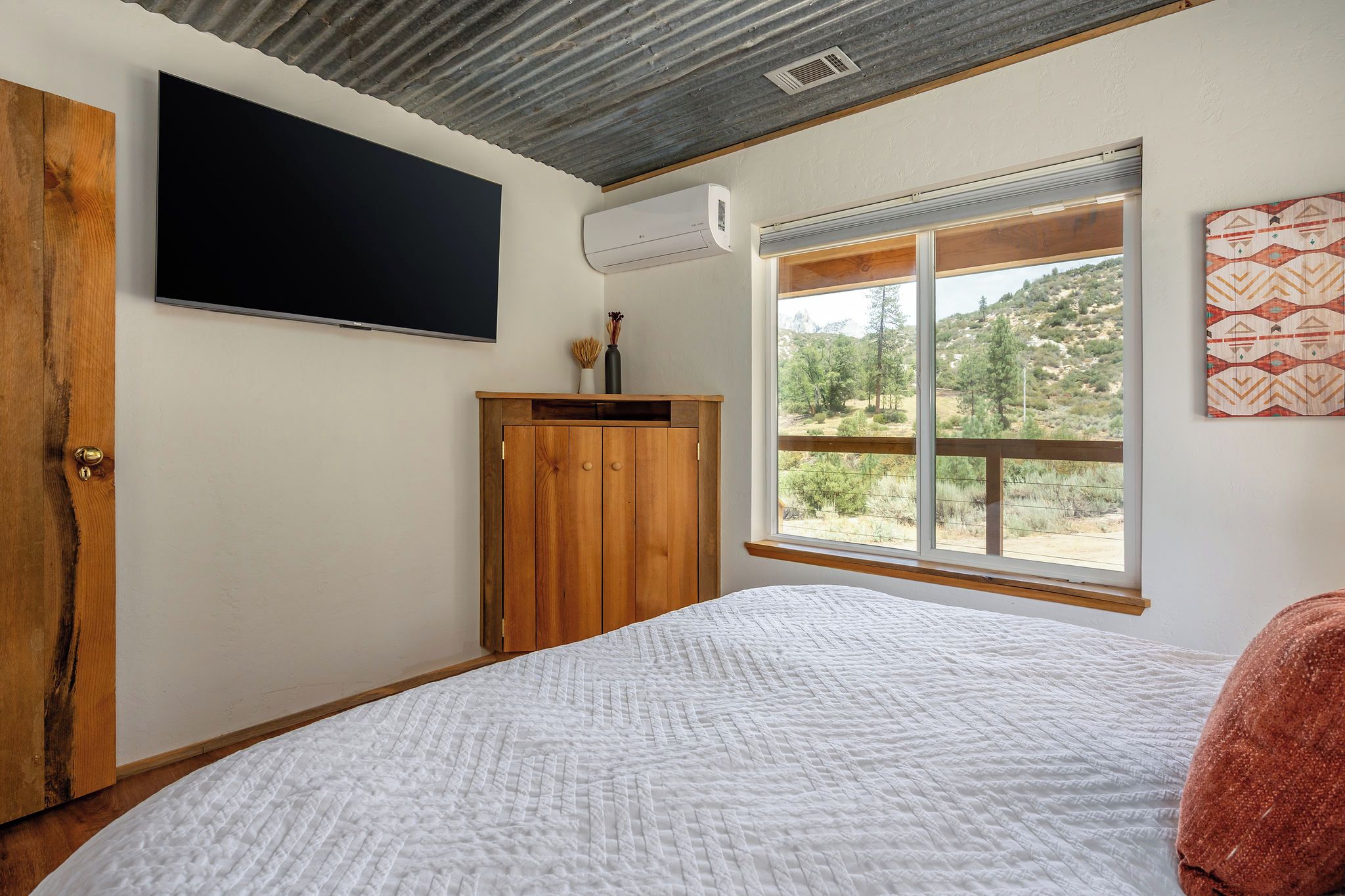 A bedroom with a bed, a wooden wardrobe, a flat-screen TV, an air conditioning unit, and a window with a view of trees.