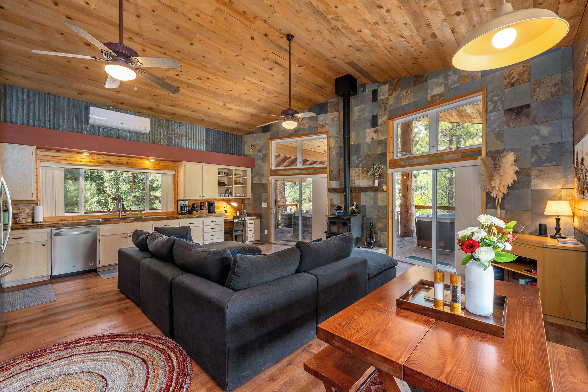 A modern interior with a wooden ceiling, a black sofa, a wooden dining table, and a kitchen area with a stone backsplash.