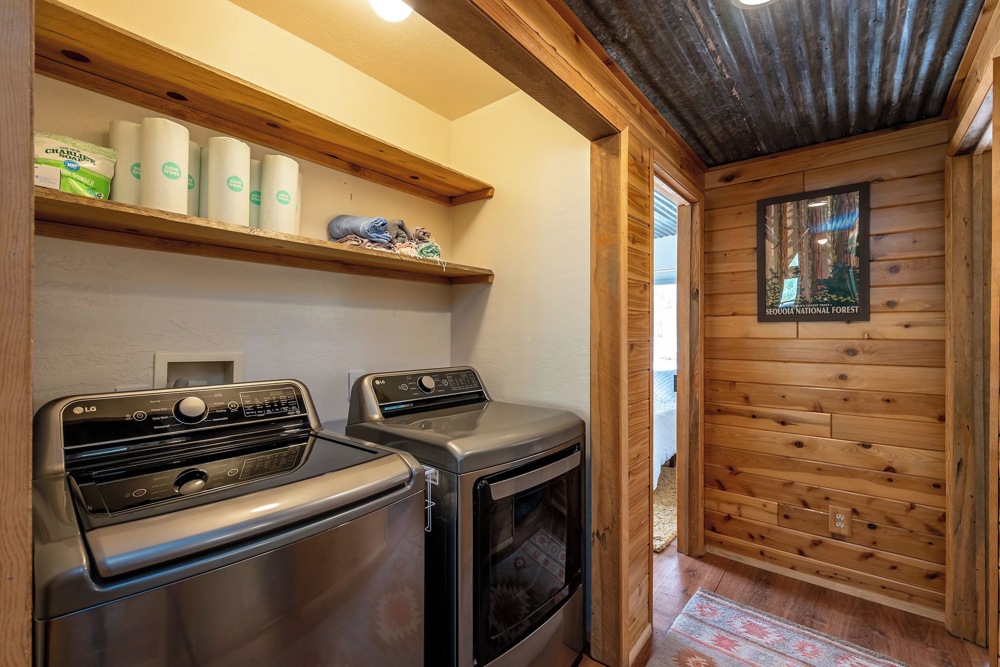 A laundry area with two LG washing machines, a shelf with paper towels, and a framed picture of a forest.