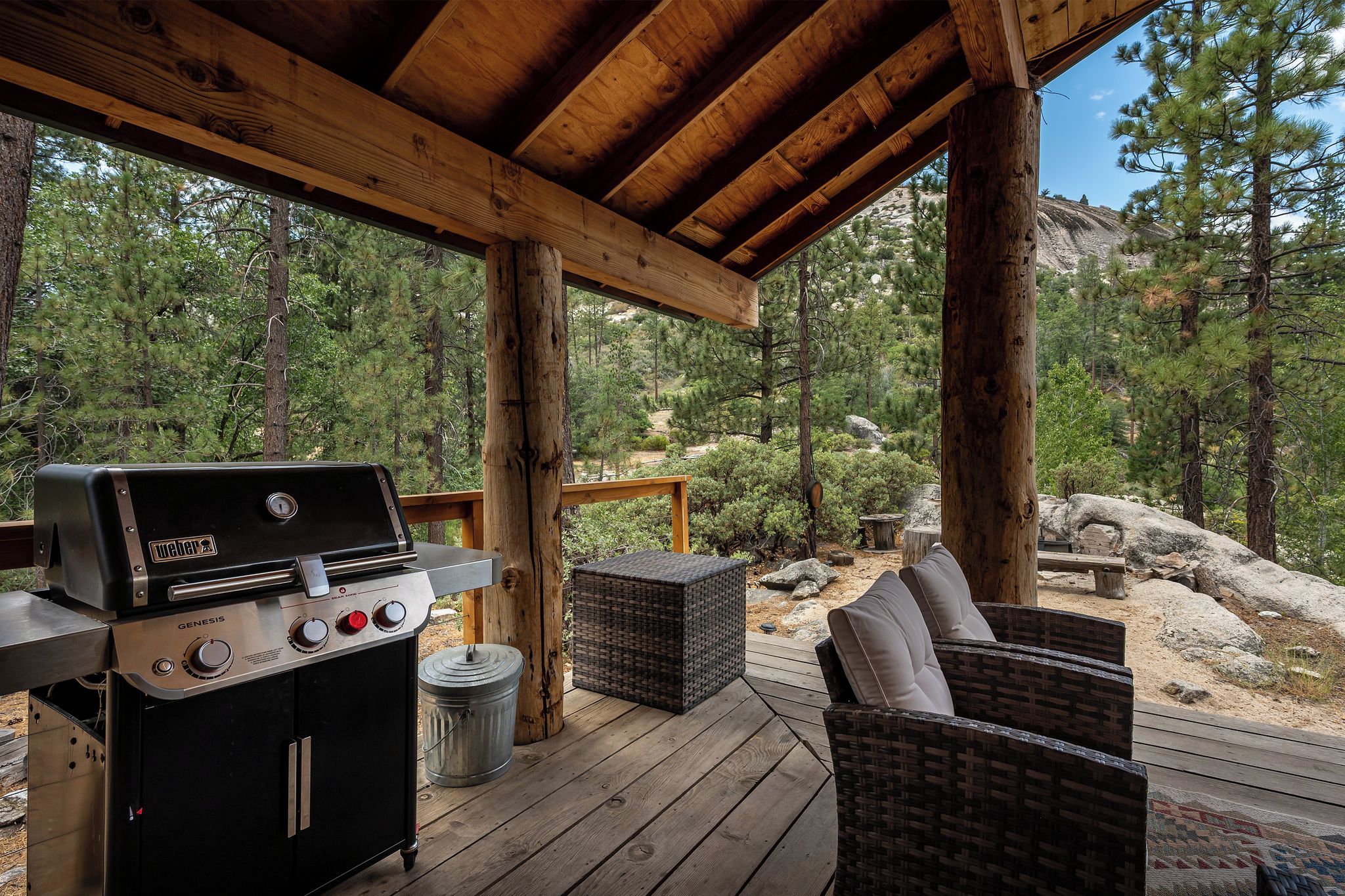 A covered outdoor area with a grill, wicker furniture, and a view of a forested landscape.