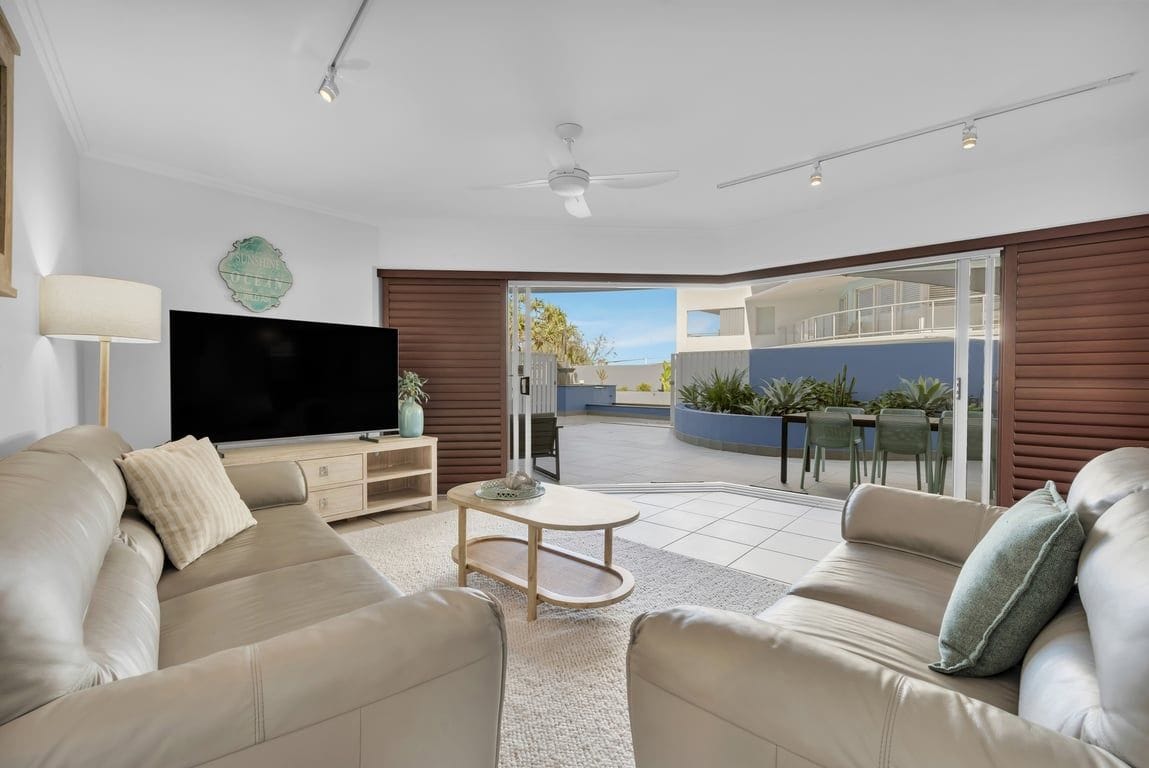 A modern living room with a flat-screen TV, leather sofas, a coffee table, and a sliding glass door leading to an outdoor patio.