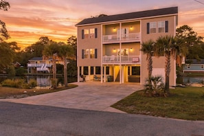 A two-story building with a balcony, a covered front porch, and a driveway leading to the entrance. - Banana Villa - Tropical 4BR Pond-Front Home w/ Private Hot Tub (Walk to Beach) (North Myrtle Beach)