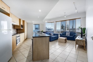 A modern kitchen with a central island, a dining area with a blue sofa, and large windows offering a city view. - 2 Bedroom Poolside Rooftop No Balcony (Kings Beach)