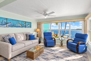 Sala con vista al mar, ventilador de techo y sofá con cojines azules.