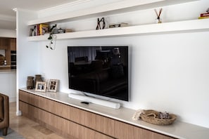 Ein modernes Wohnzimmer mit einem Flachbildfernseher an einer weißen Wand, Holzschränken darunter und einem Regal mit Dekorationselementen.