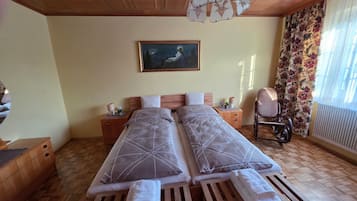 A double bed with a geometric patterned bedspread, a bedside table with a lamp, a painting on the wall, and a window with floral curtains.