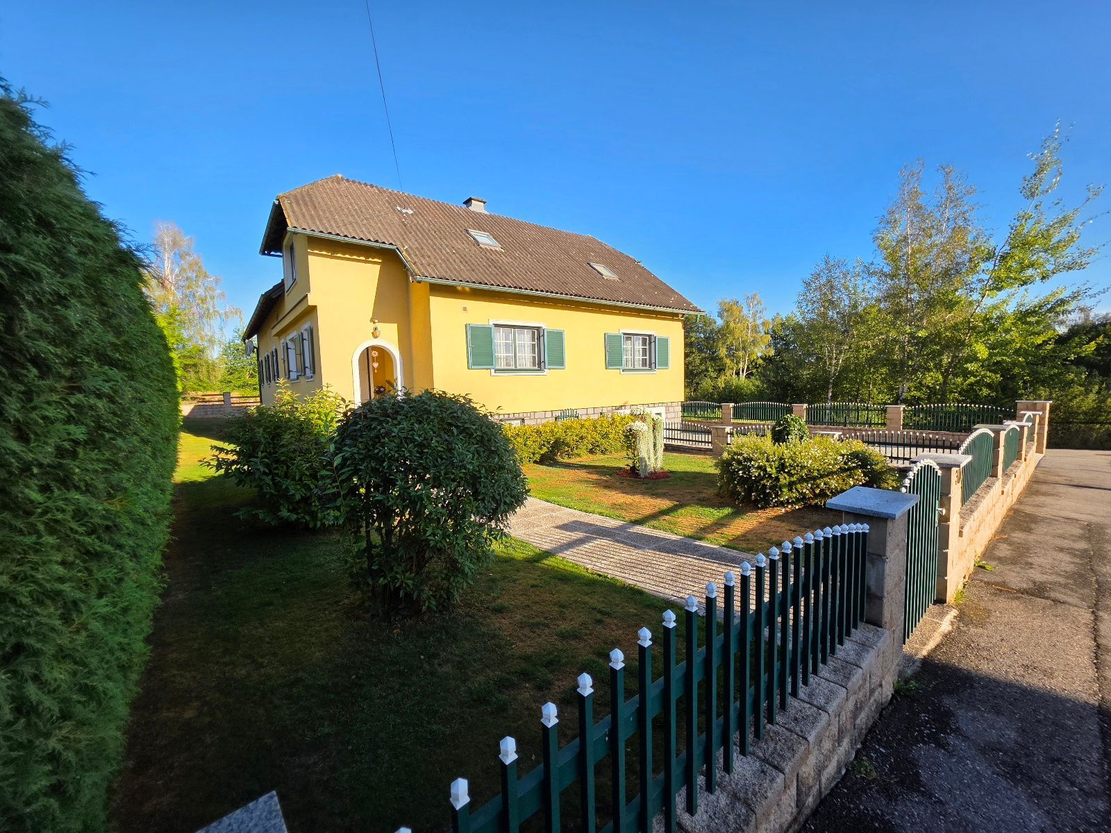 Ett gult hus med ett grönt staket, en asfalterad uppfart och en välskött trädgård.