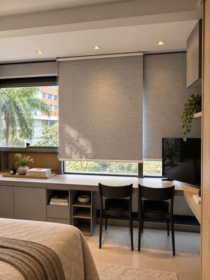 A modern bedroom with a large bed, a desk with two chairs, a TV, and a window with a view of palm trees. - Studio apartment in Pinheiros | Near Oscar Freire Metro Station, swimming pool and gym. (São Paulo)