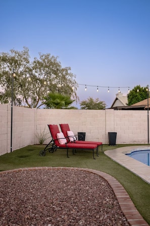 A backyard with a swimming pool, red lounge chairs, and a string of lights. - Private Pool | Game Room | Mesa Family Retreat (Mesa)