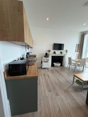 A modern kitchen with a microwave, a coffee maker, and a small cabinet. There is a dining area with a table and chairs, and a fireplace with a TV mounted above it. - Seaside 2BR Stay in Herne Bay (Herne Bay)