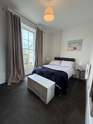 Een slaapkamer met een bed, nachtlampen, een raam met gordijnen, een nachtkastje en een aan de muur hangend kunstwerk.