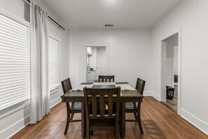 A dining area with a wooden table and chairs, a kitchen in the background, and a laundry basket.