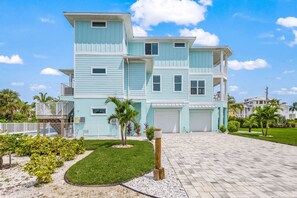 A multi-story residential building with a light blue exterior, multiple garage doors, and a paved driveway. - Beachside Bliss with Heated Pool, Spa, Bikes & Views - Cozy Cottage - Roelens (Fort Myers Beach)