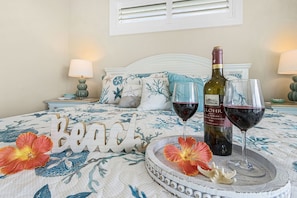 A bedroom with a bed, a nightstand, a bottle of wine, two wine glasses, and a decorative word 'Beach' on the bed. - Beachside Bliss with Heated Pool, Spa, Bikes & Views - Cozy Cottage - Roelens (Fort Myers Beach)