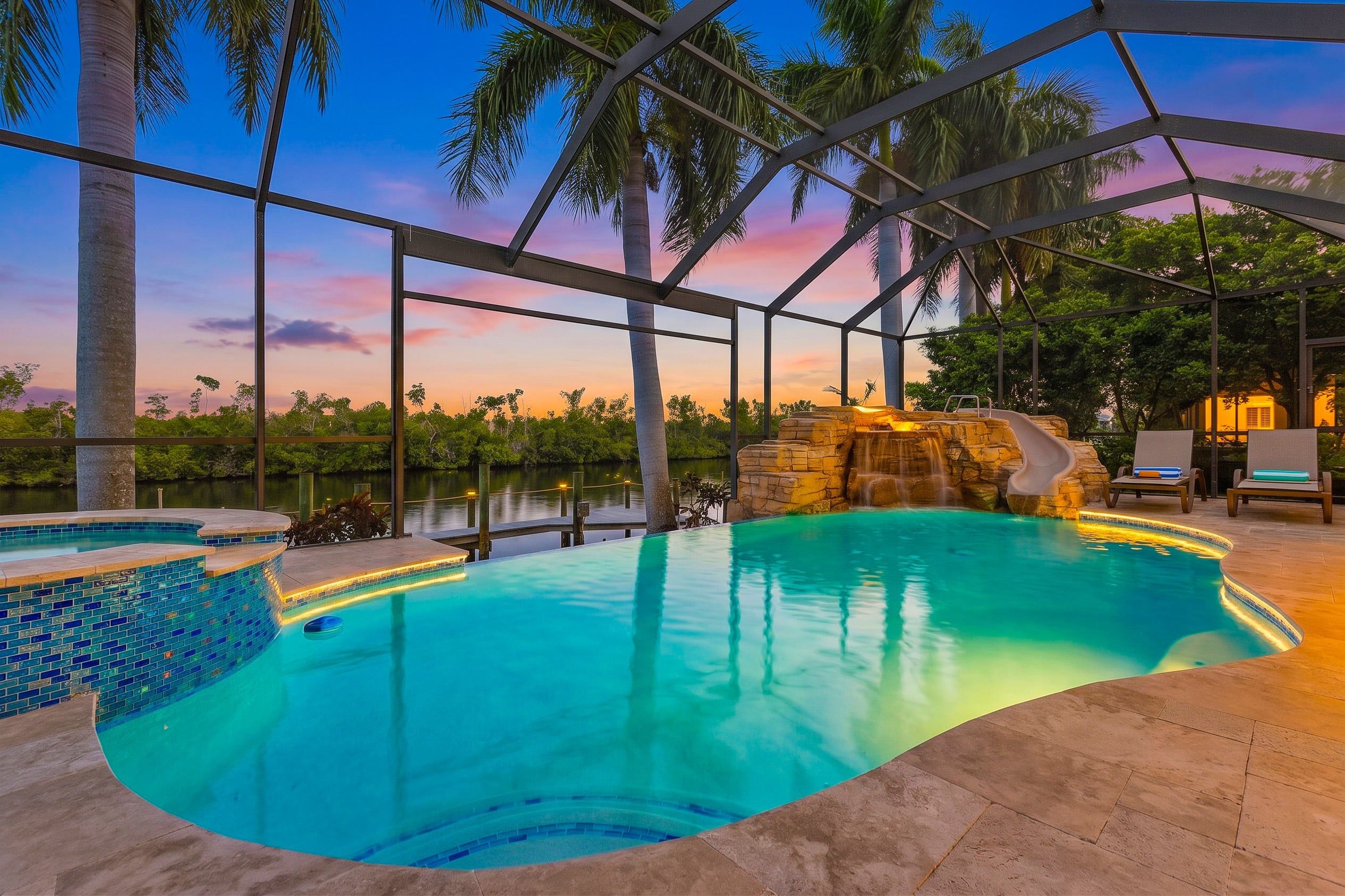 An indoor swimming pool with a view of the sunset, surrounded by palm trees and a stone waterfall feature.
