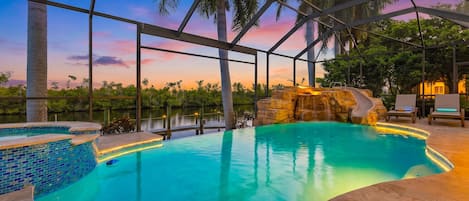 An indoor swimming pool with a view of the sunset, surrounded by palm trees and a stone waterfall feature.