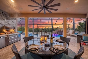 An outdoor dining area with a glass table, wicker chairs, and a view of the sunset. - Waterfront Fun, Pool & Spa Paradise with Game Room & Gulf Access - Palm Vista Villa - Roelens (Cape Coral)