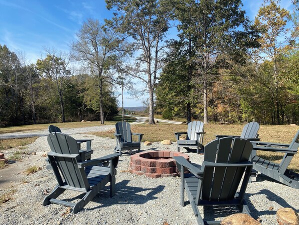A gravel area with Adirondack chairs and a fire pit, surrounded by trees and a clear sky.