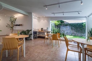 A modern dining area with a kitchen in the background, a large window with a view of greenery, and a ceiling with recessed lighting. - Herbolaria Hotel (San Salvador)