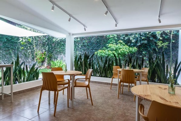 An indoor dining area with tables and chairs, large windows, and a view of lush greenery outside. - Herbolaria Hotel (San Salvador)