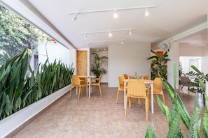 Espaço moderno em ambiente interno com área de refeições, mesas e cadeiras de madeira, cercado por grandes plantas verdes.