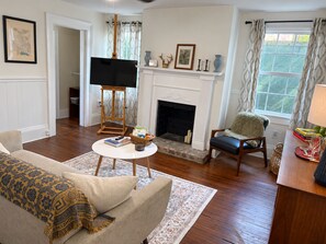 Una sala de estar con chimenea, un televisor sobre un mueble bajo, un sofá, una silla y una mesa con libros y una cesta.