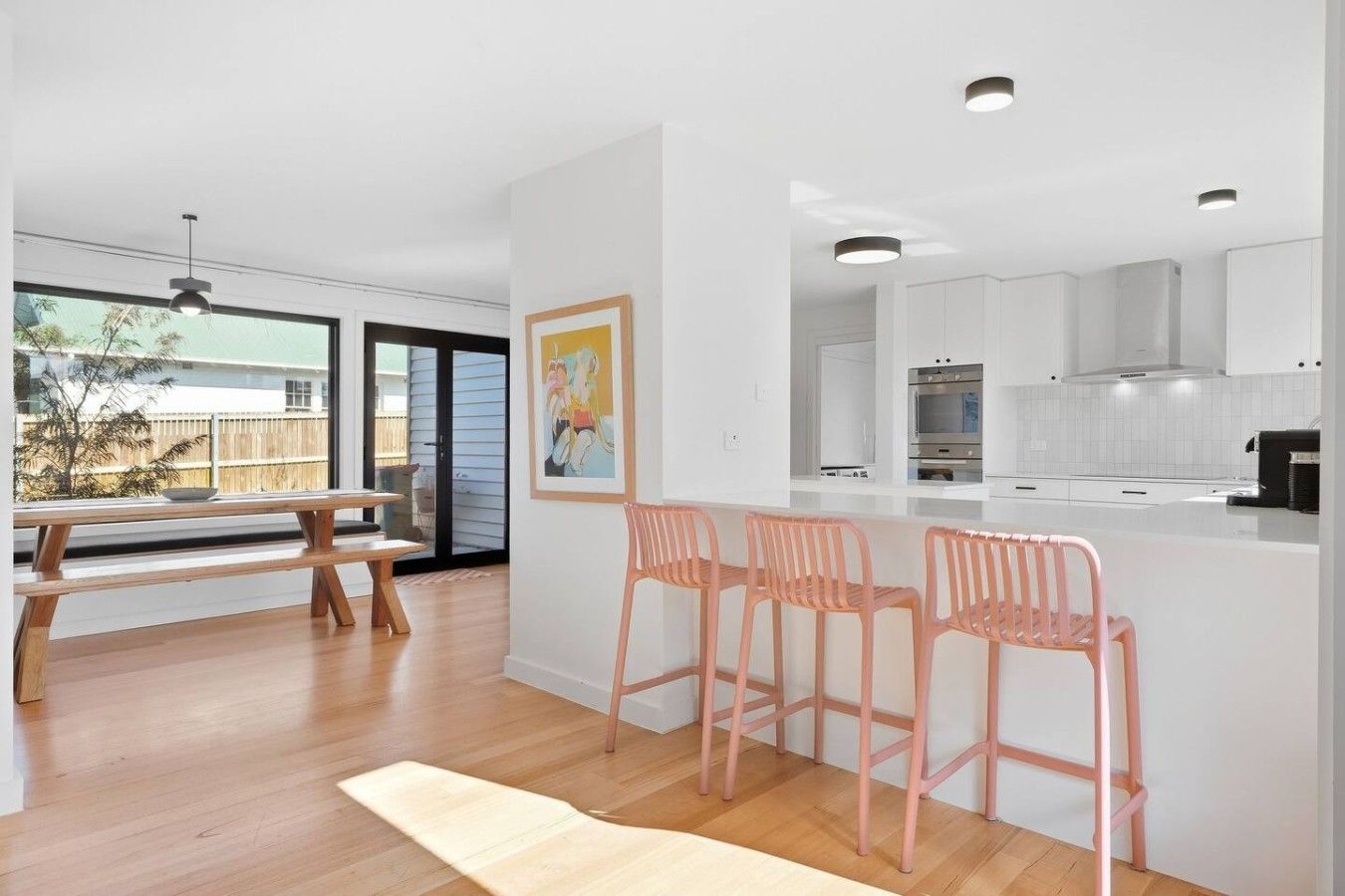 A modern kitchen with white cabinets, a stainless steel oven, and a countertop. There are three pink chairs at a wooden table with a light-colored rug.