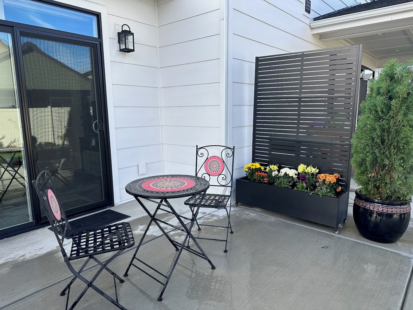 Une terrasse avec une table et deux chaises, un jardinier avec des fleurs et un arbre en pot.