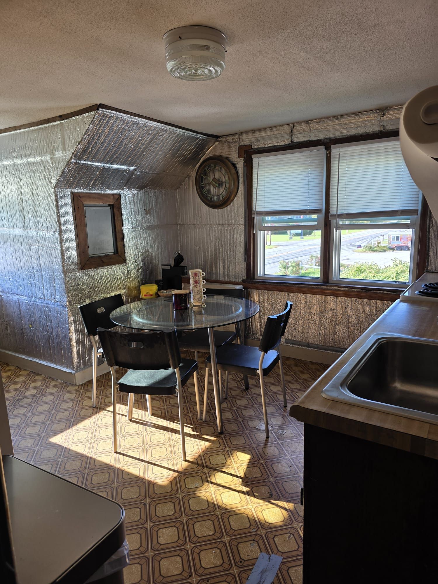 A kitchen with a round glass table, two chairs, a clock, and a window with blinds.