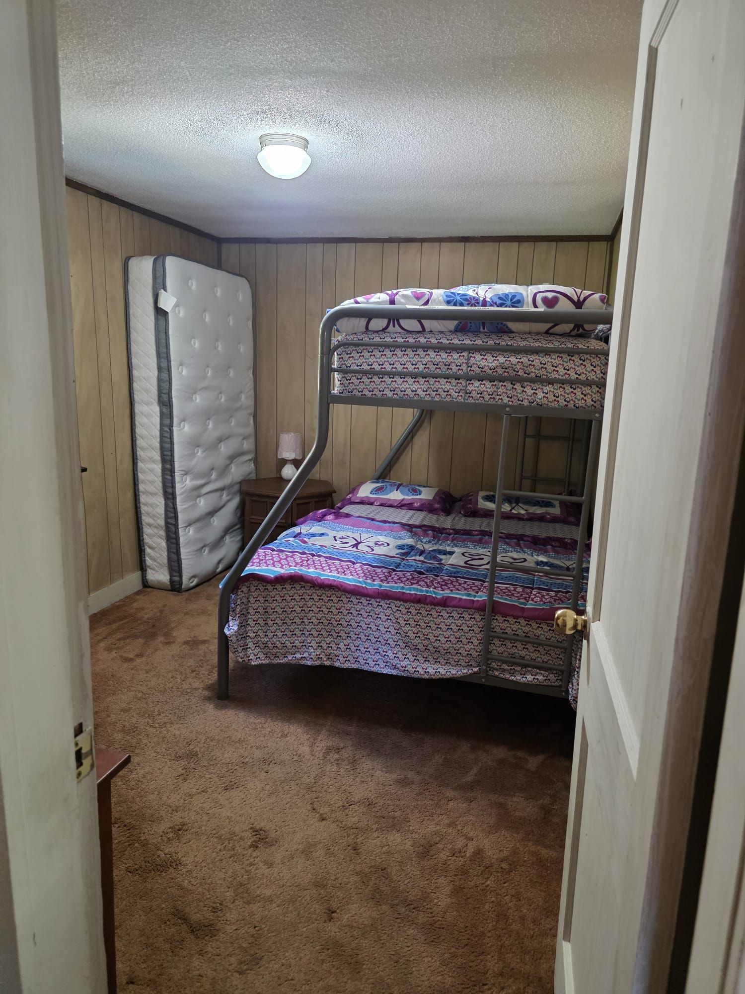 A bunk bed with patterned bedding, a single mattress against the wall, and a carpeted floor.