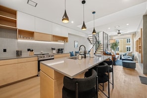 A modern kitchen with a central island, pendant lighting, and a staircase leading to a living area with seating. - 3-Story Designer Townhome by City Park (Manhattan)